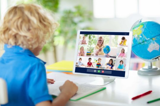 Young boy working at a table listening to a video lesson with his teacher and classmates.