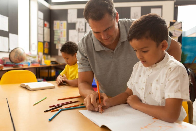 Male teacher helping a young boy at his desk with colored pencils.