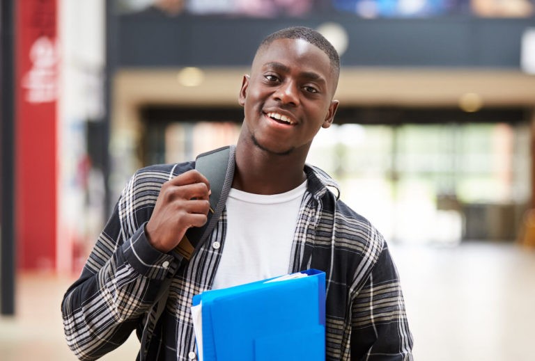 Young man standing with a backpack and a binder in a college hallway.