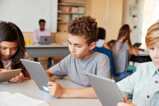 Young students sitting at a table in a classroom using tablets.