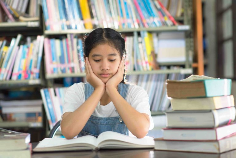 Young girl sitting at a library table with books looking bored with her hands on her face