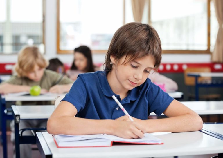 Young boy sitting at a desk in a classroom writing in a journal.