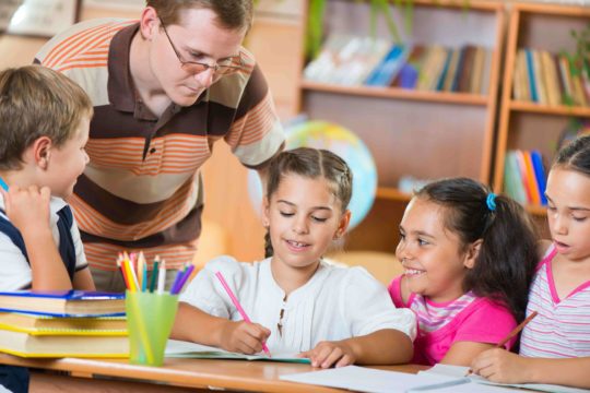 Male teacher hovering over table with young students while they draw