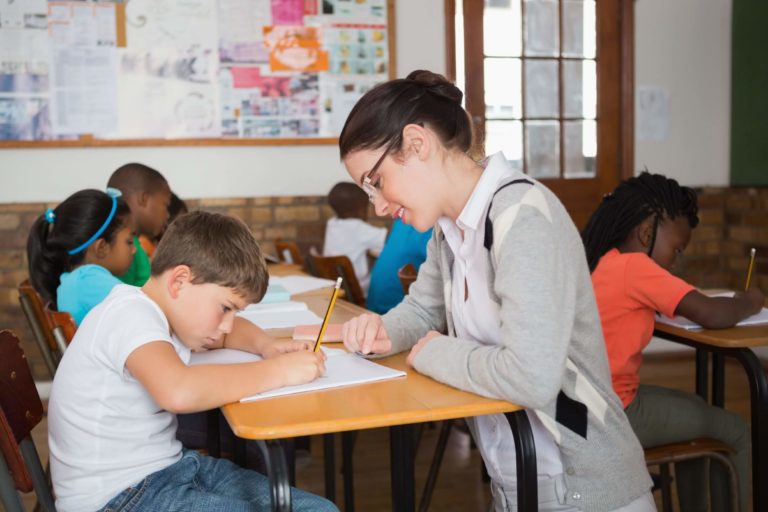 Female teacher helping a young boy at his desk in a classroom.