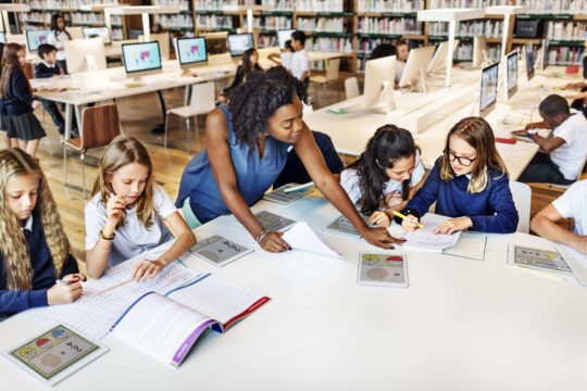 Female teacher helping students at a table working on fractions on tablets