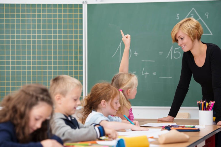 Teacher standing by a group of students with a girl raising her hand.