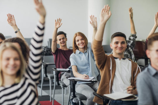 Classroom of high school students smiling and holding up their hands.