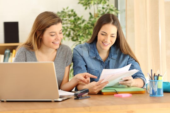 Two young ladies sitting at a table looking at a paper together.