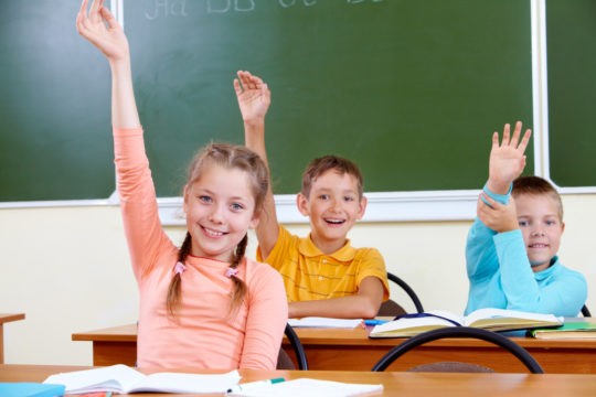 Young smiling students sitting at desks with books open in front of them while raising their hands