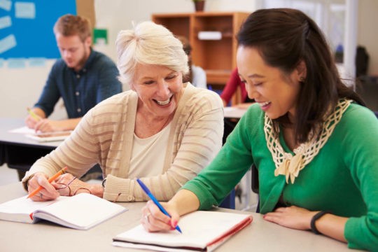 Older teacher sitting at a table with a younger teacher sharing notes.