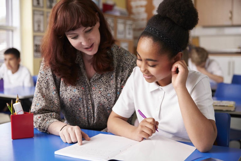 Teacher helping a young student writing at her desk.