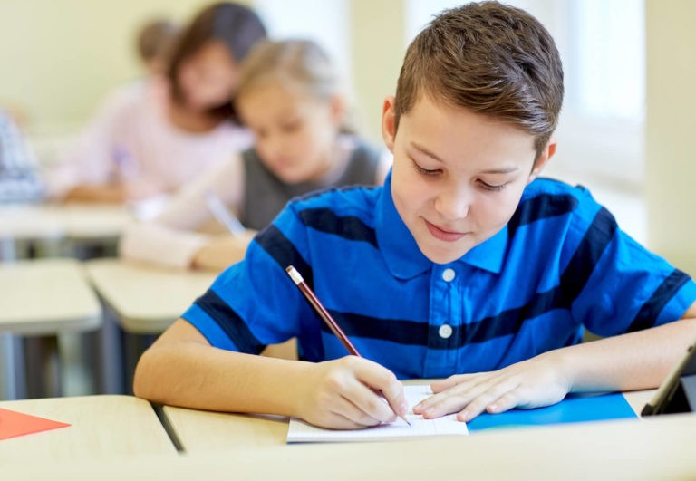 Content looking boy sitting at his desk writing in a classroom with students.