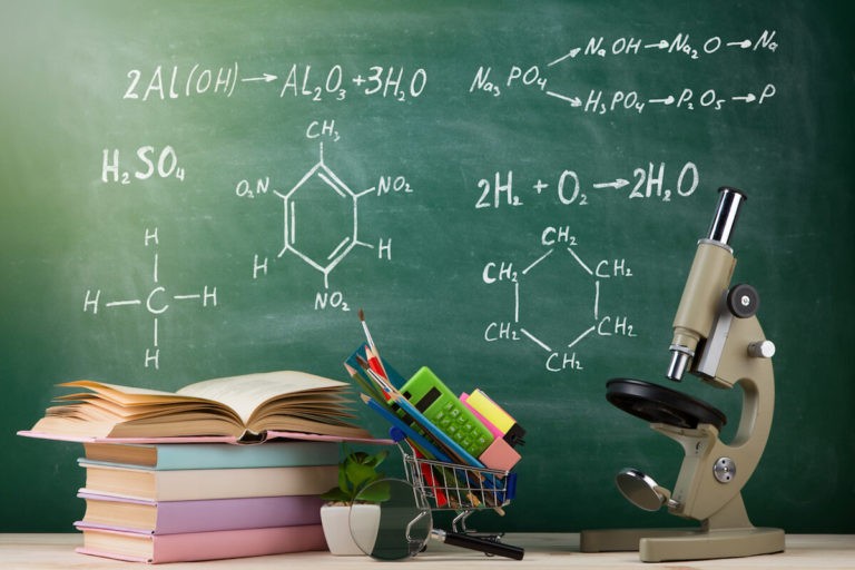 A group of books, a microscope, and calculators sit on a teacher’s desk in front of a chalkboard with science equations written out on it.