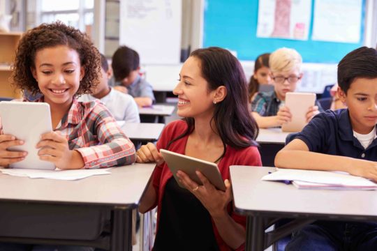 Smiling teacher holding a tablet and helping a student at her desk.