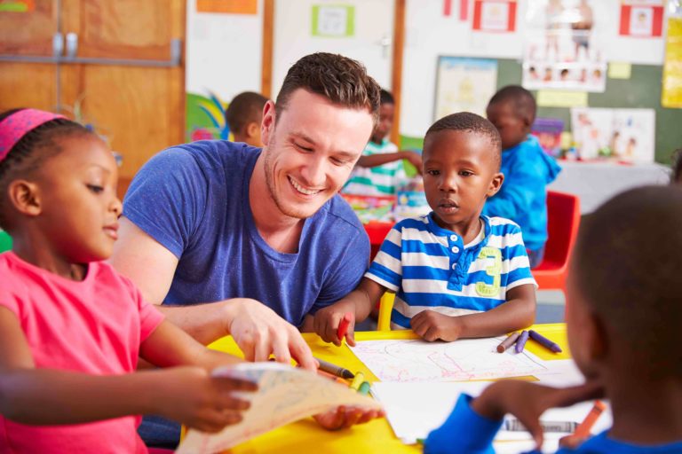 Male teacher sitting at table drawing with young children