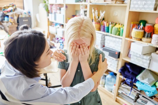 Teacher comforting a crying girl standing in an art classroom.