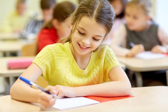 Young girl sitting at a desk in a classroom writing on paper.